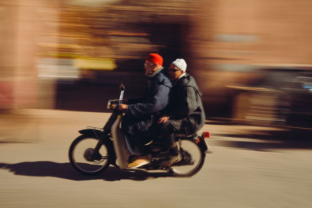 Two people wearing winter hats ride a motorcycle through a city street, captured with motion blur effect.