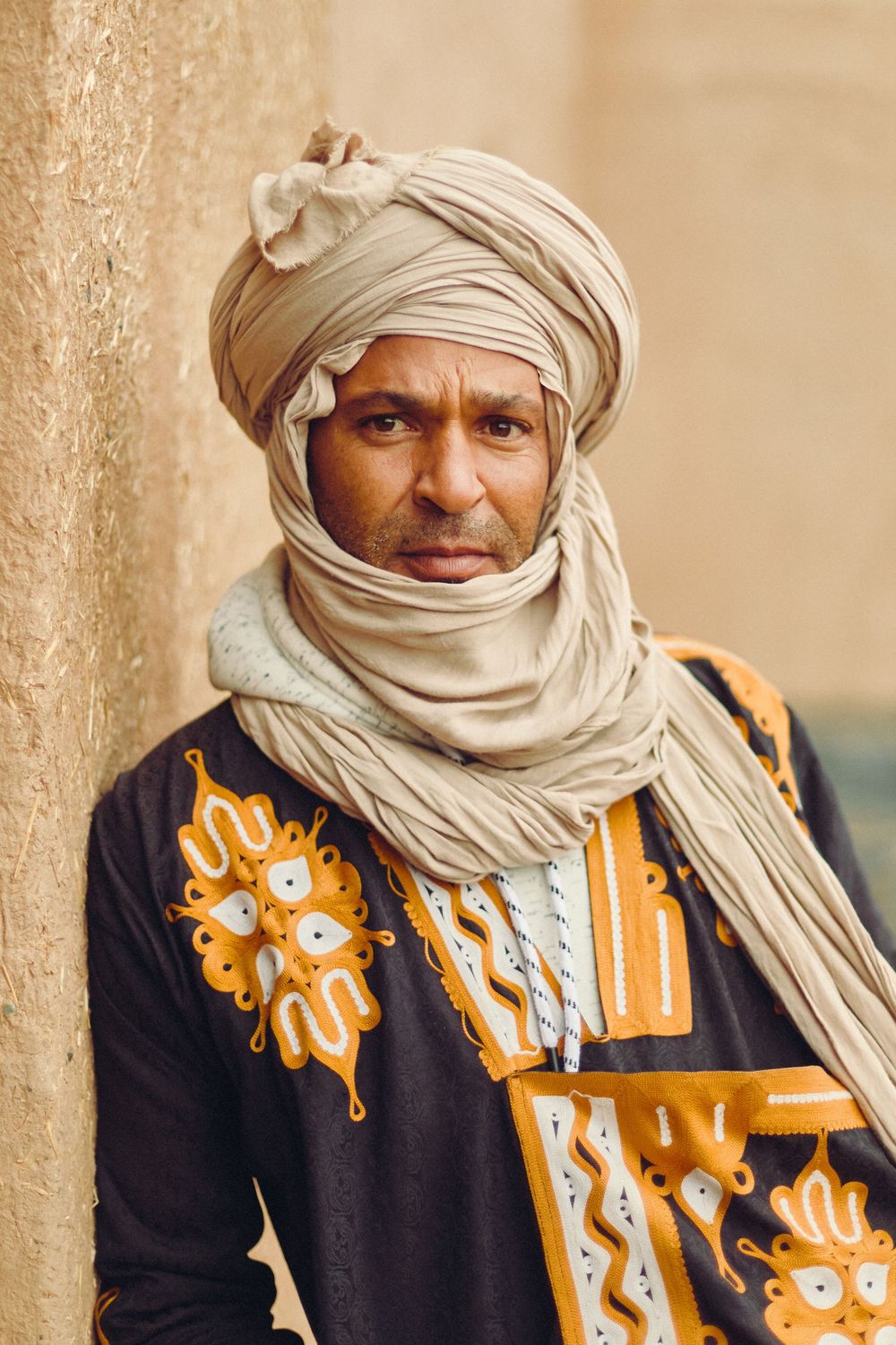 A Tuareg man in traditional embroidered robe and beige turban poses against a sandy wall.