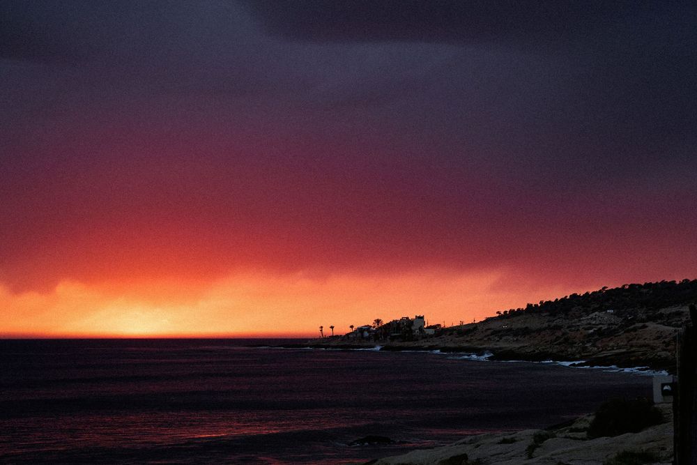 Dramatic coastal sunset with vivid red and orange sky over dark stormy clouds above a calm ocean shoreline.