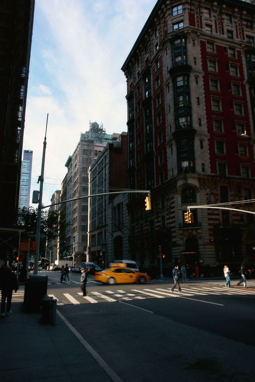 Yellow taxi cab crossing a busy New York City intersection with pedestrians, traffic lights, and classic brick buildings.