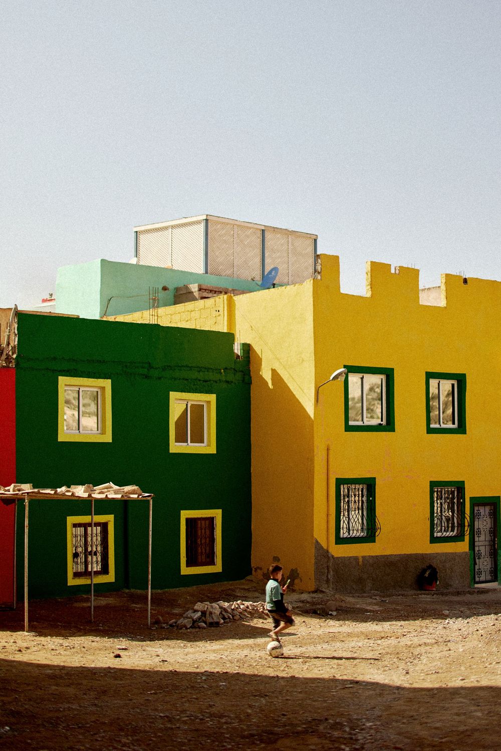 Colorful green and yellow buildings with a child walking in a sandy courtyard under a clear sky.