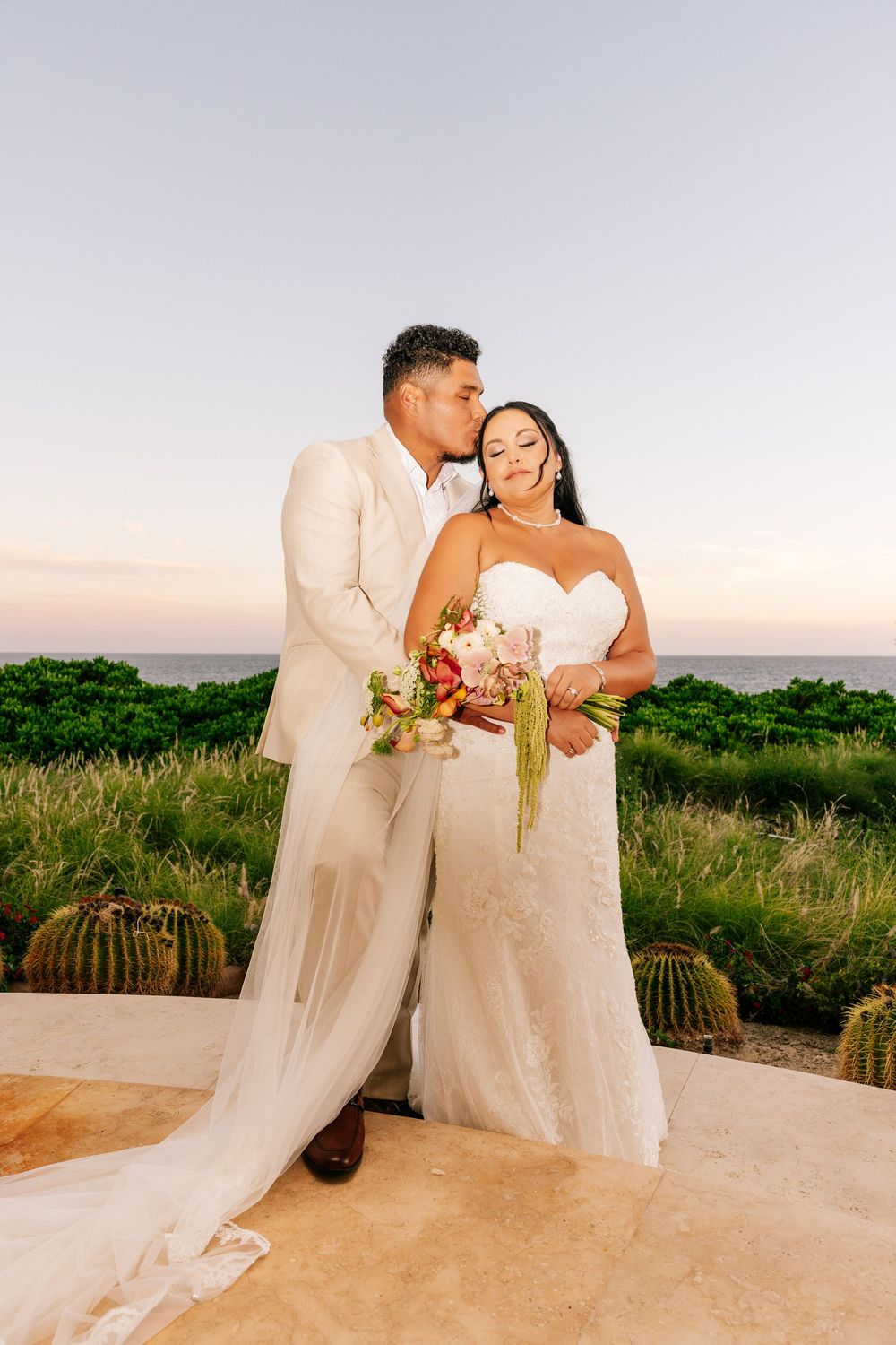 A groom in a cream suit kisses his bride on the forehead as she holds a bouquet on a terrace overlooking the ocean at sunset.