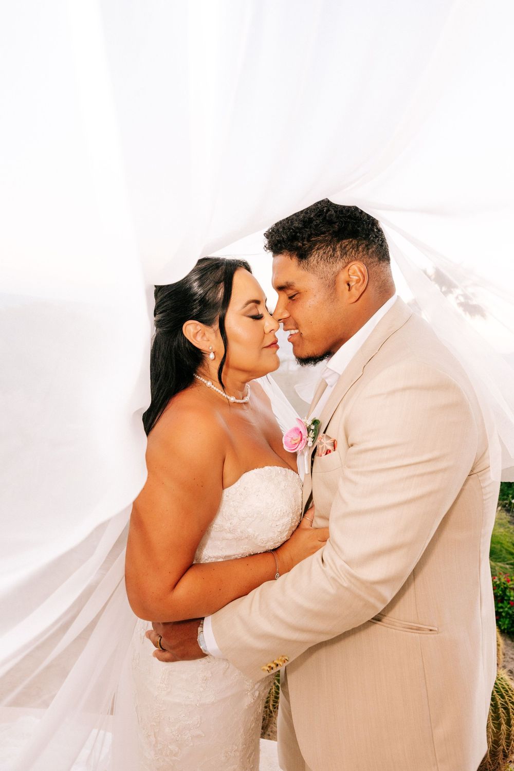Bride in a white strapless lace gown and groom in a tan suit with a pink boutonniere share a tender moment beneath a flowing white veil.