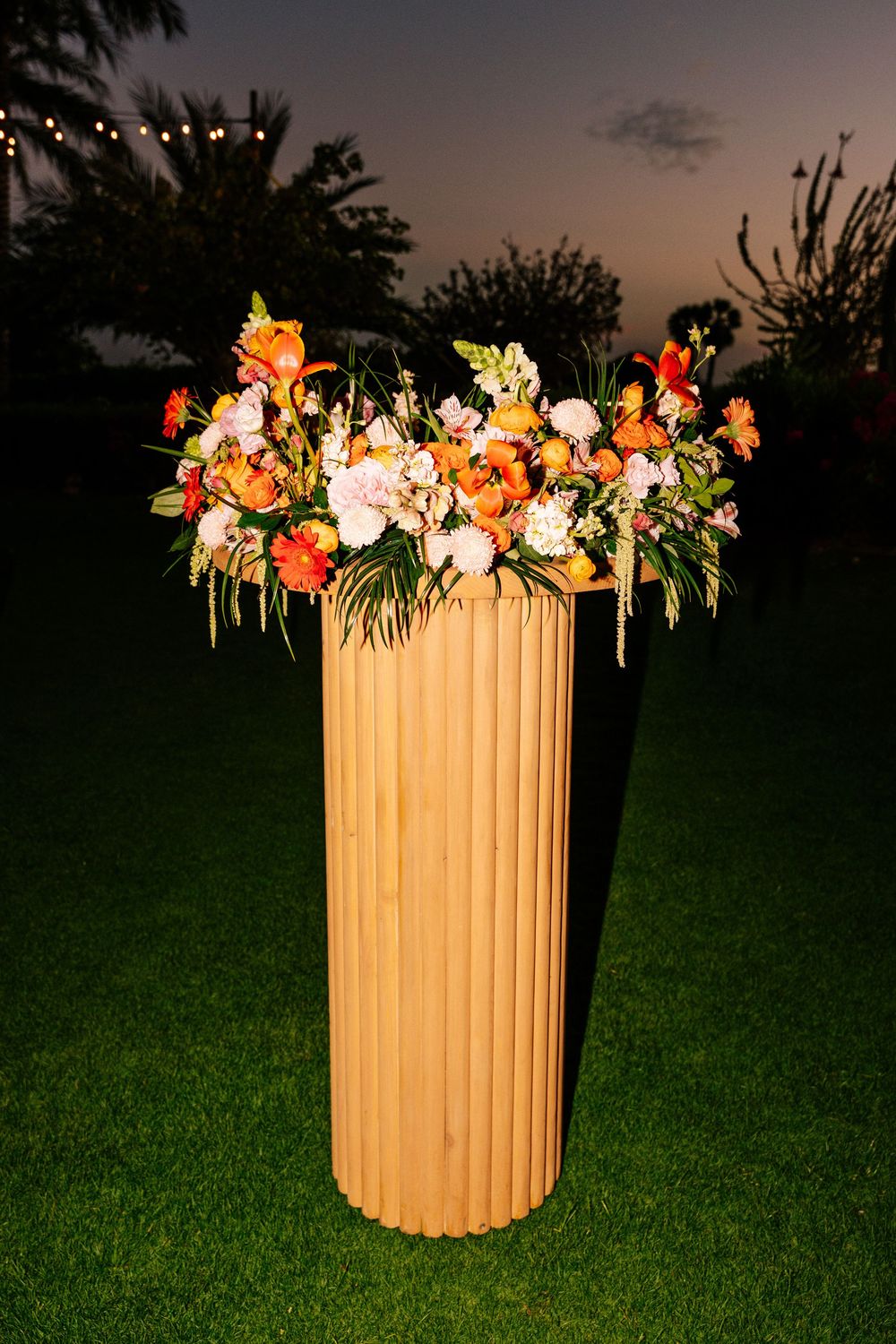 Orange, red and white flower arrangement atop a tall wooden fluted pedestal on a lawn at dusk with string lights in the background.