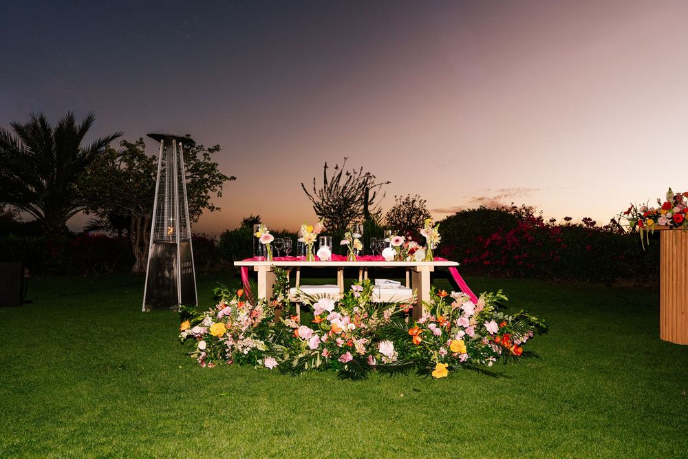 Pink-trimmed table surrounded by colorful flower arrangements on a green lawn at dusk, with a patio heater and palm trees nearby.