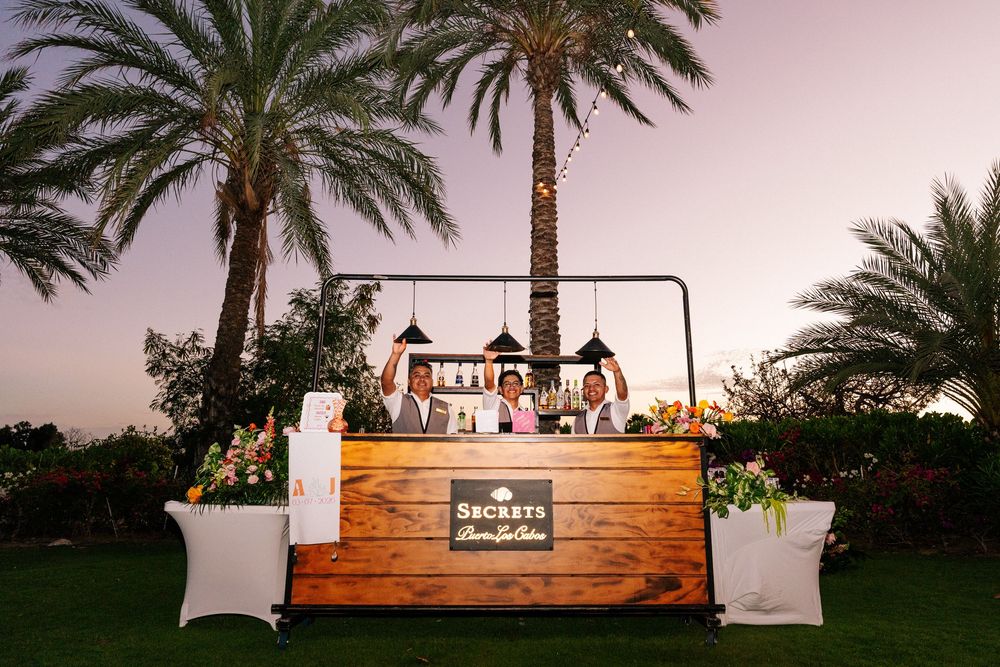 Three bartenders stand behind a rustic wooden Secrets bar with floral accents on a lawn surrounded by palm trees at sunset.