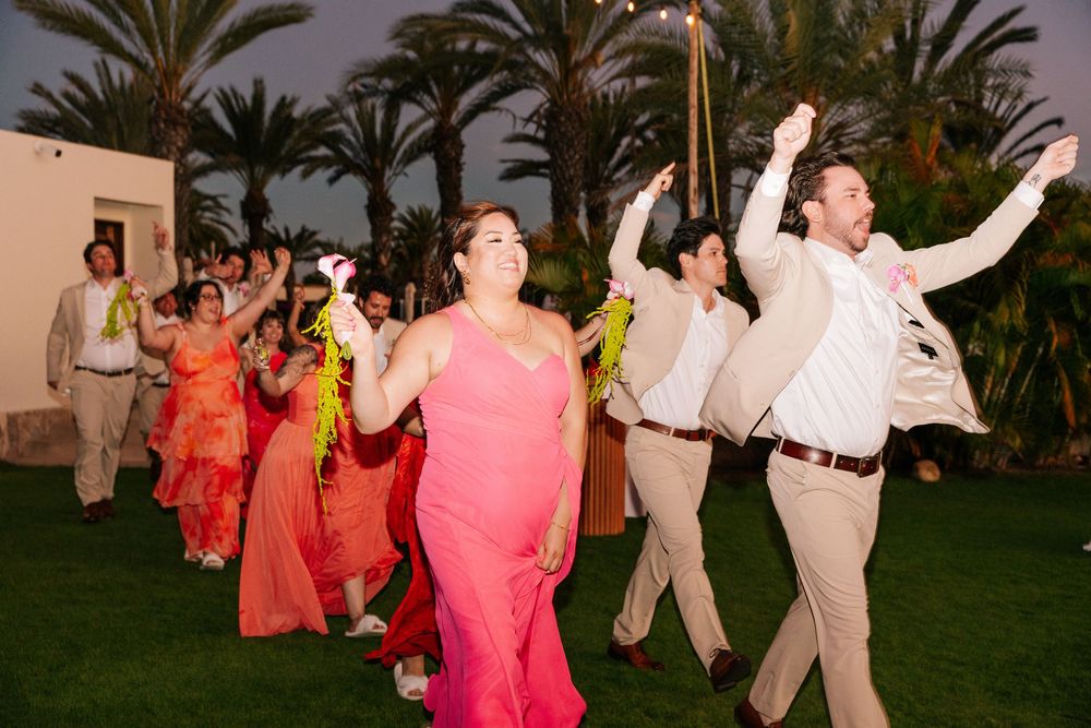 Wedding party in coral dresses and tan suits dances joyfully on a lawn among palm trees at a tropical evening celebration.