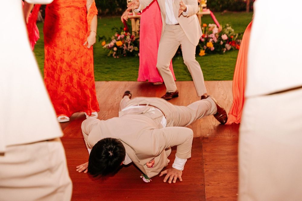 Groomsman in a tan suit bows low to the ground on a wooden dance floor at an outdoor wedding as guests in formal attire look on.