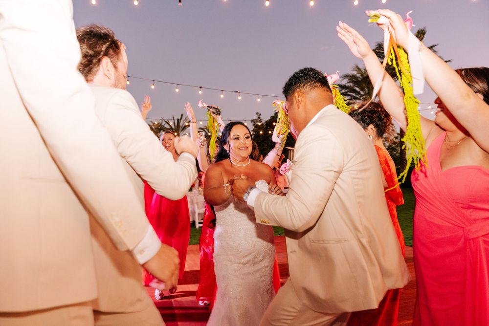 Bride in a white gown dances with a groom in a tan suit as guests in coral dresses cheer at an outdoor evening wedding reception.