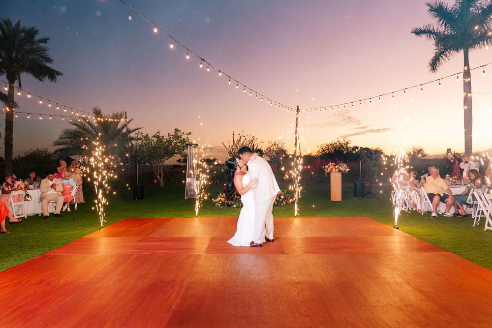 Bride and groom share a first dance on a wooden dance floor at a tropical sunset wedding with string lights, palm trees and seated guests.