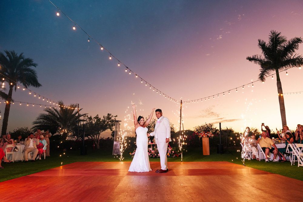 Bride and groom share a first dance on a wooden dance floor under string lights at sunset, with palm trees and guests watching nearby.