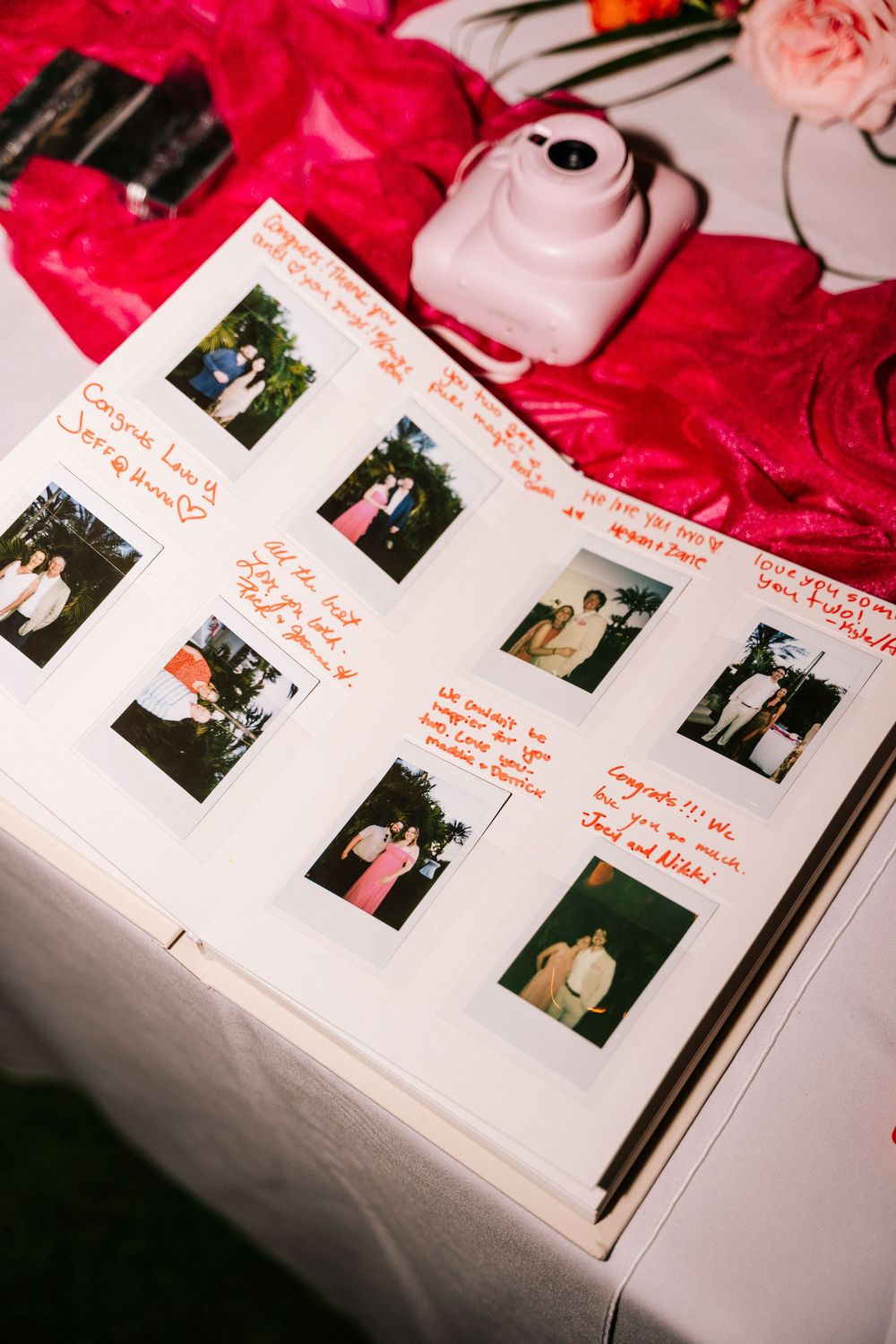 Open wedding guest book with Polaroid photos and handwritten orange notes, beside a pink instant camera on a table with red linens.