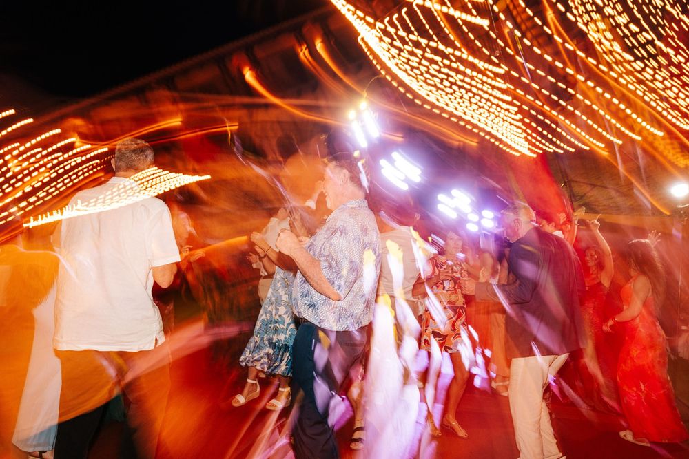 Guests dance under glowing string lights at an evening outdoor party, with motion blur capturing the lively energy of the celebration.