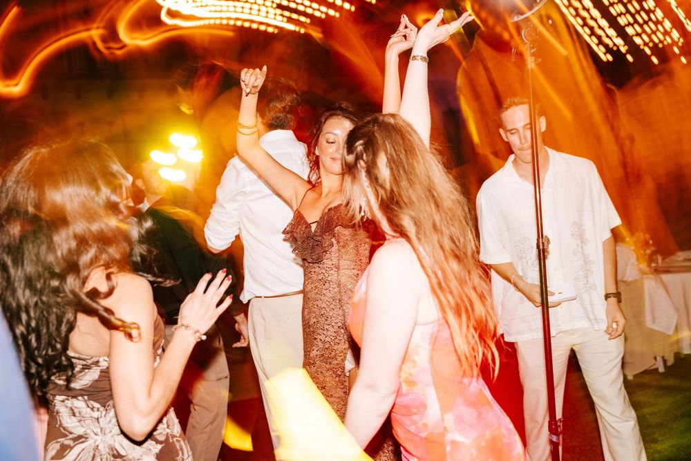 Women in party dresses dance and raise their arms joyfully on a lively dance floor while a man in white looks on during an evening event.
