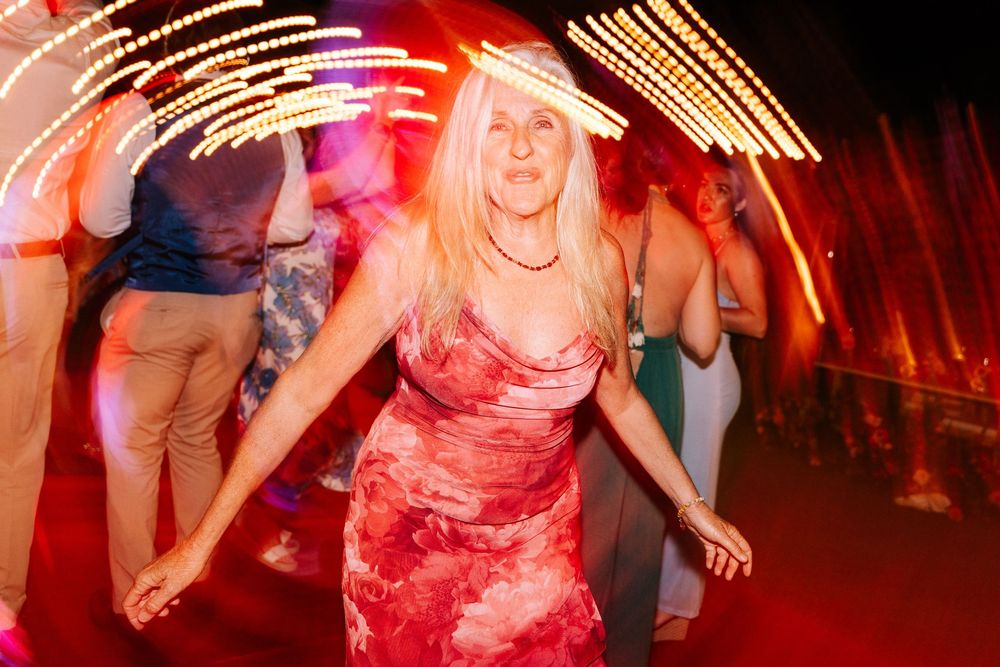 A woman in a red floral dress dances joyfully with arms outstretched as glowing lights blur around guests at an evening wedding reception.