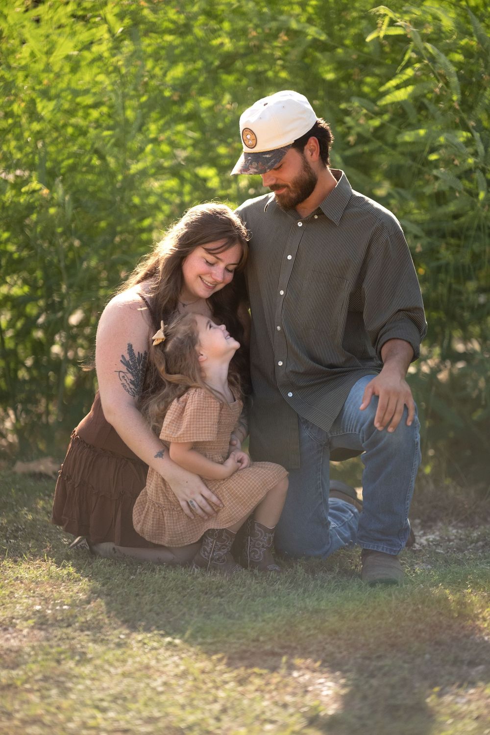A young family poses outdoors in golden light, parents kneeling beside their toddler daughter in a lush green setting.