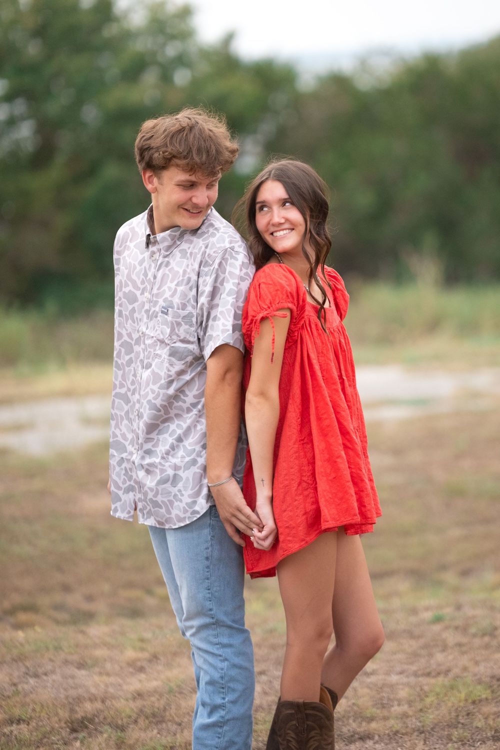 A young couple holds hands and smiles at each other outdoors, she in a red dress and boots, he in a patterned shirt.