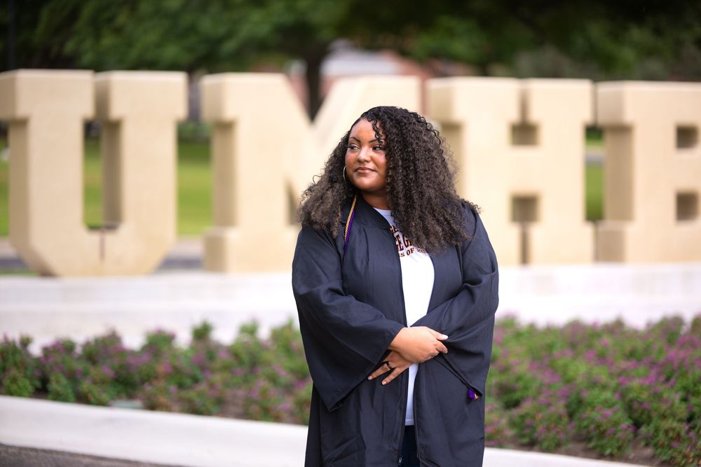 Graduate in cap and gown poses proudly in front of large university letters outdoors.