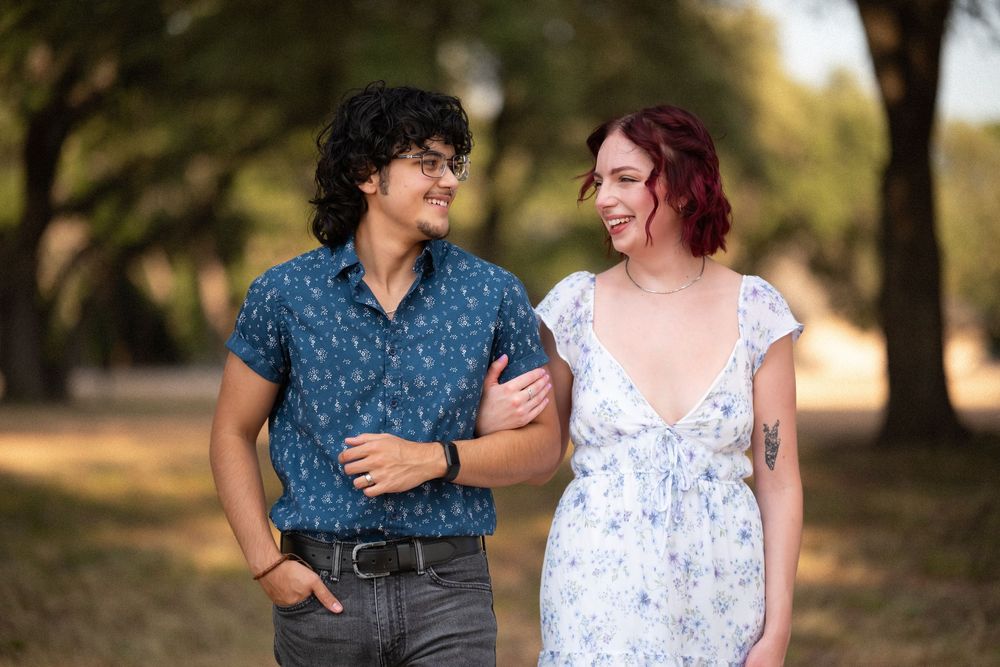 A smiling couple walks arm in arm through a sunlit park, the woman in a floral dress and the man in a blue patterned shirt.