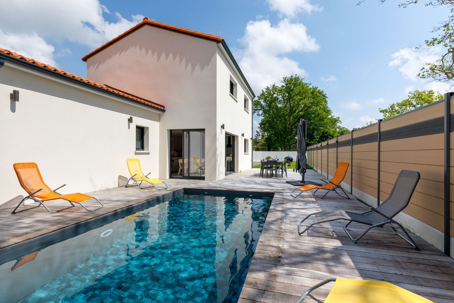Villa moderne avec piscine privée, terrasse en bois et chaises longues orange par une journée ensoleillée.