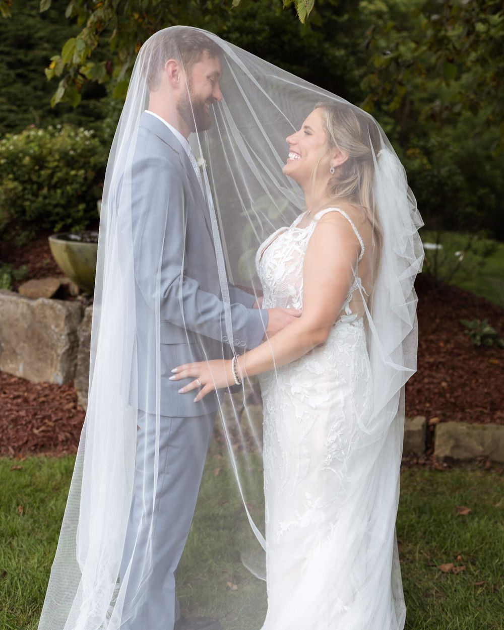 Smiling bride in a lace gown and groom in a gray suit share a tender moment beneath a sheer wedding veil in a lush garden.