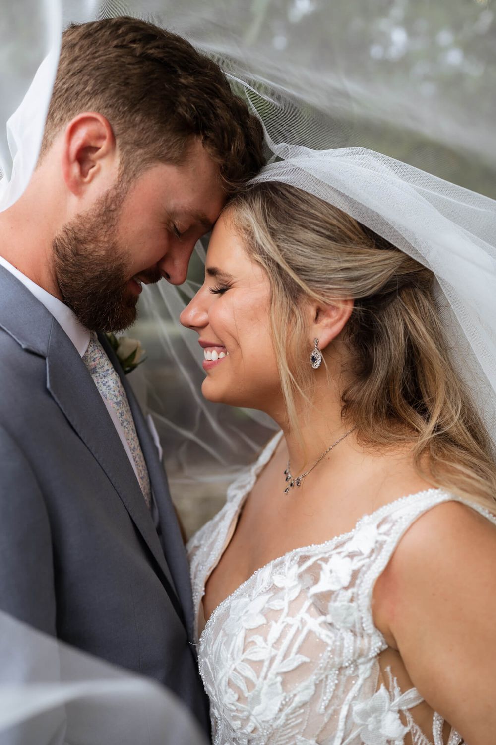 Smiling bride in a lace wedding gown and veil touches foreheads with her bearded groom in a gray suit during a tender wedding portrait.