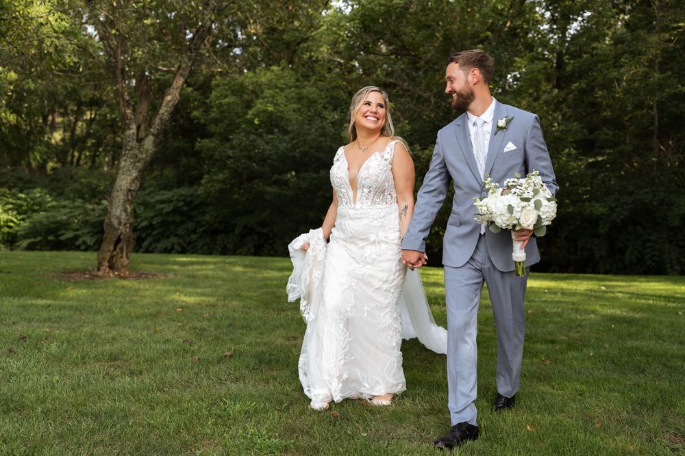 Smiling bride in a lace wedding gown and groom in a gray suit holding a white bouquet walk hand in hand across a sunny green lawn.