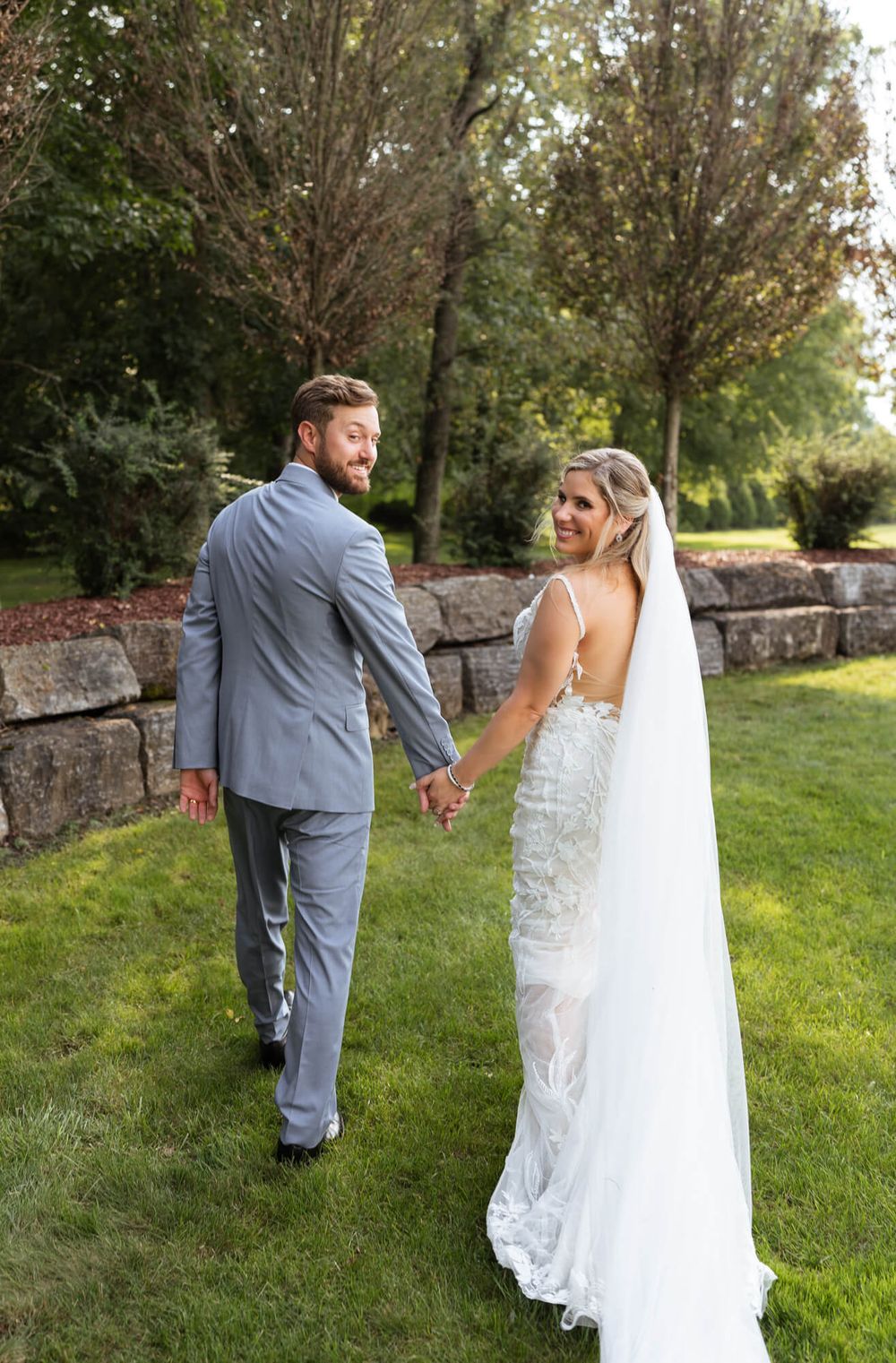 Bride in a lace wedding gown and long veil holds hands with her groom in a gray suit while walking on a lawn by a stone wall.