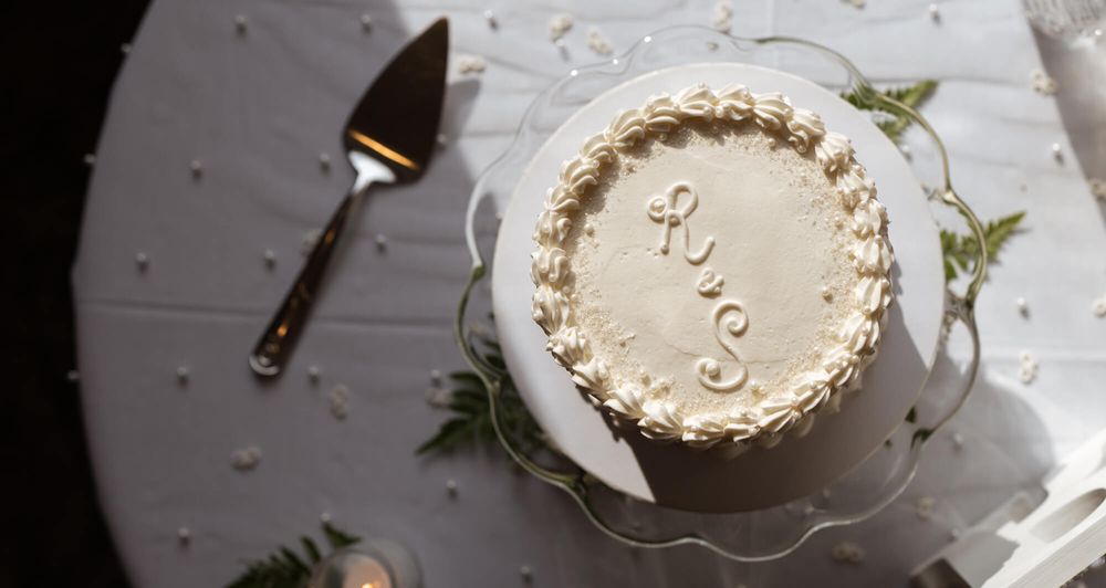Small round wedding cake with white buttercream piping and a monogram, displayed on a glass stand beside a silver cake server.