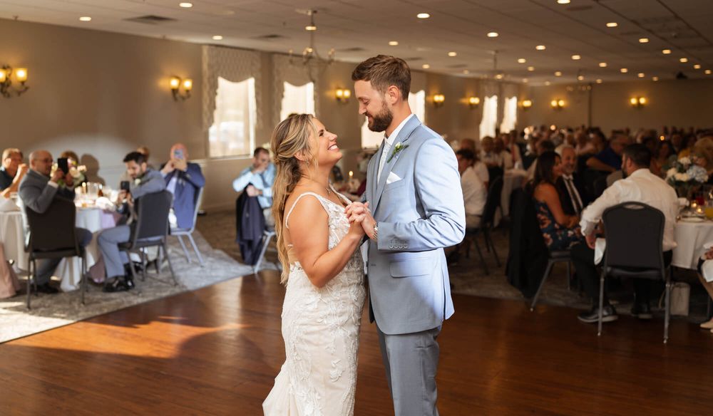 Bride in a white gown and groom in a light gray suit share a first dance on a wood floor while wedding guests watch from tables.