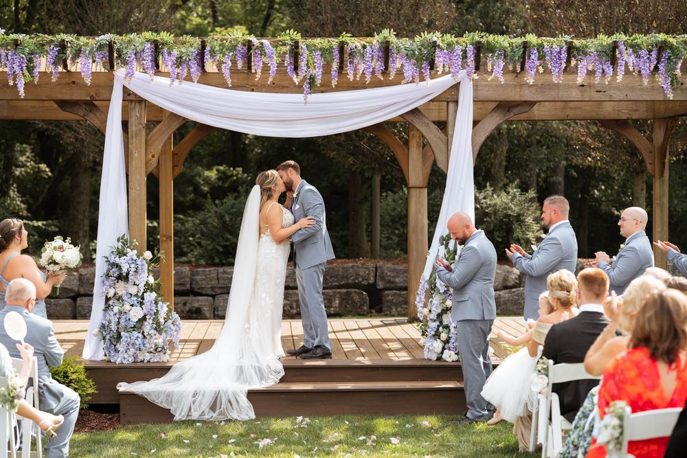 Bride and groom exchange vows under a wooden pavilion draped with white fabric and purple wisteria as guests and groomsmen watch outdoors.