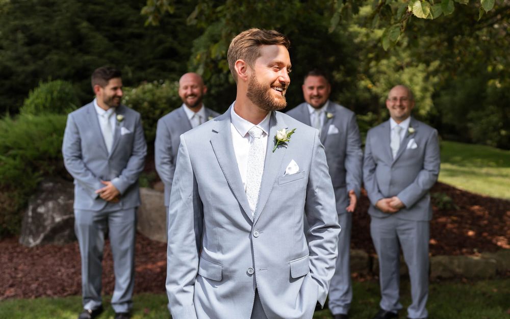 Smiling groom in a light gray suit with a boutonniere stands in a garden while four groomsmen in matching suits pose behind him.