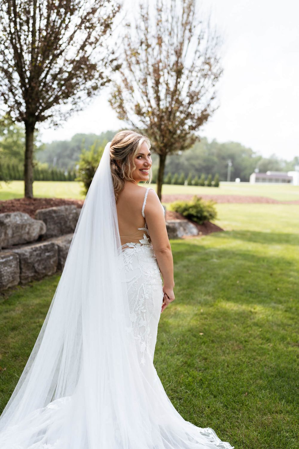 Smiling bride in a lace backless wedding gown and long cathedral veil looks over her shoulder while standing on a sunny green lawn.