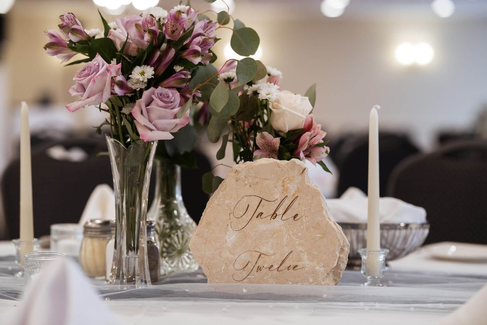 Wedding reception table with a stone number sign reading Table Twelve, mauve roses, eucalyptus in a glass vase, and white taper candles.