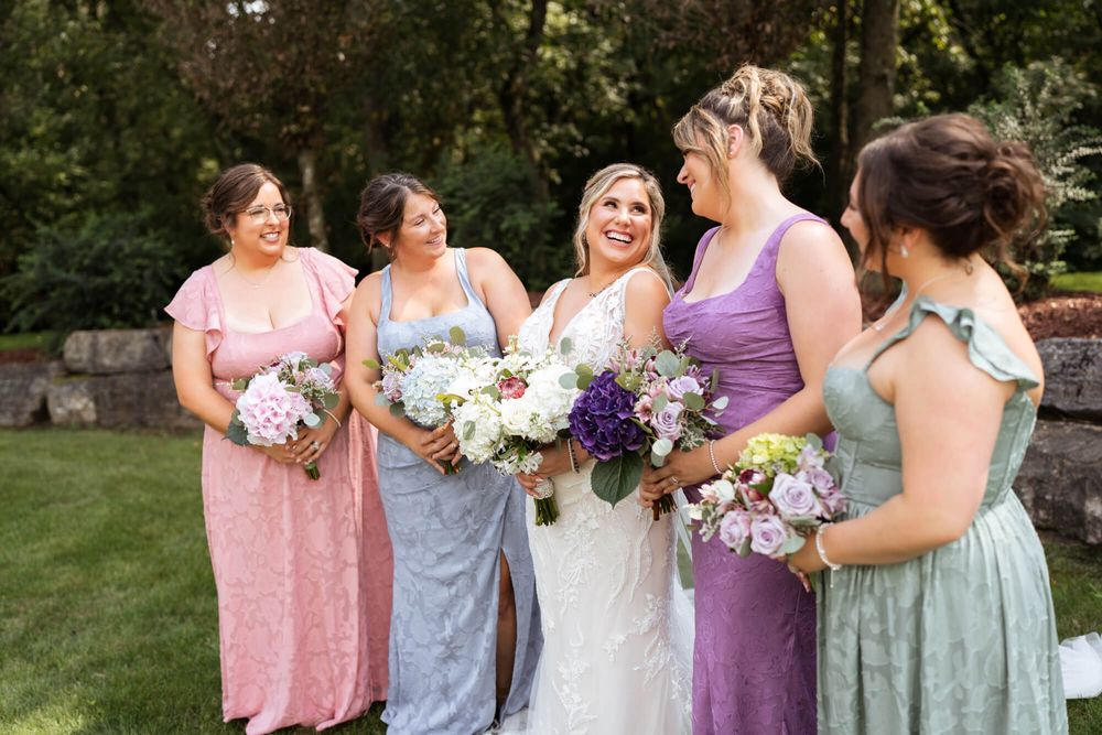 A bride in a white gown smiles among four bridesmaids in mismatched pastel dresses, all holding purple and white bouquets outdoors.