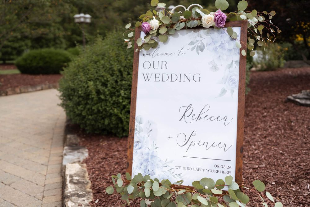 Wooden wedding welcome sign for Rebecca and Spencer decorated with pink and white flowers and greenery, set beside a garden path.