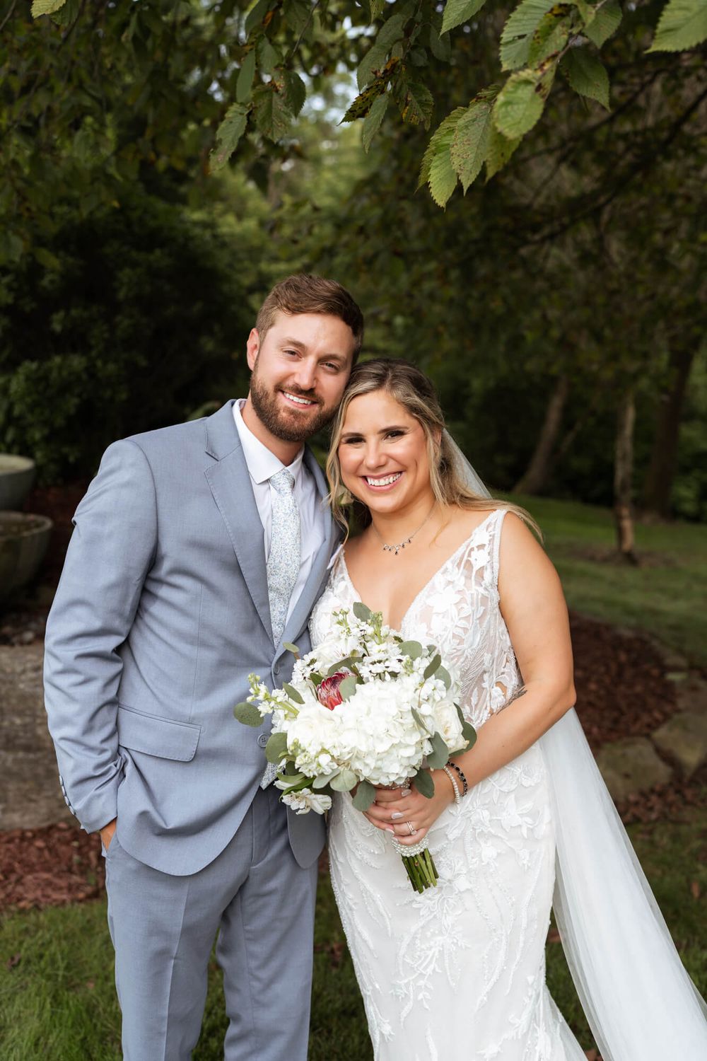 Smiling bride in a lace wedding gown and veil holds a white floral bouquet beside her groom in a gray suit in a leafy garden.