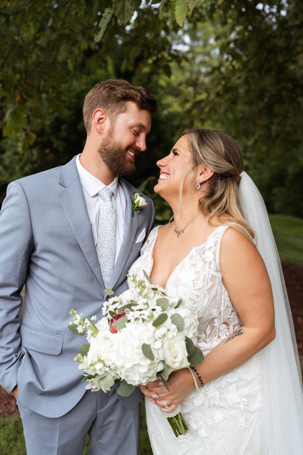 Smiling bride in a lace gown and veil holding a white floral bouquet beside her bearded groom in a light gray suit outdoors.