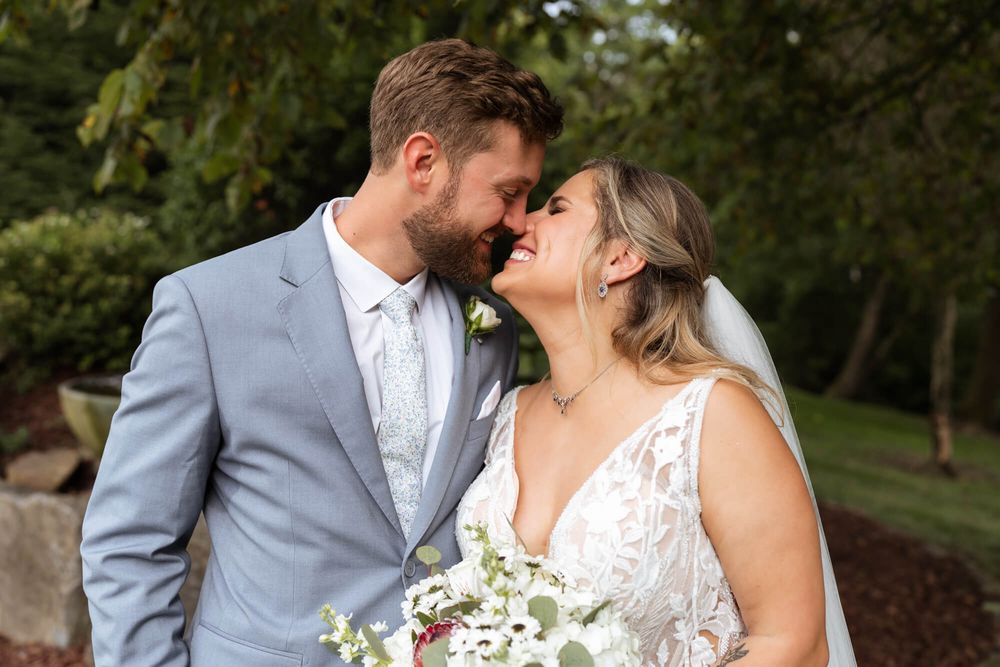 Smiling bride in a lace gown and groom in a gray suit touch foreheads while holding a white floral bouquet outdoors.