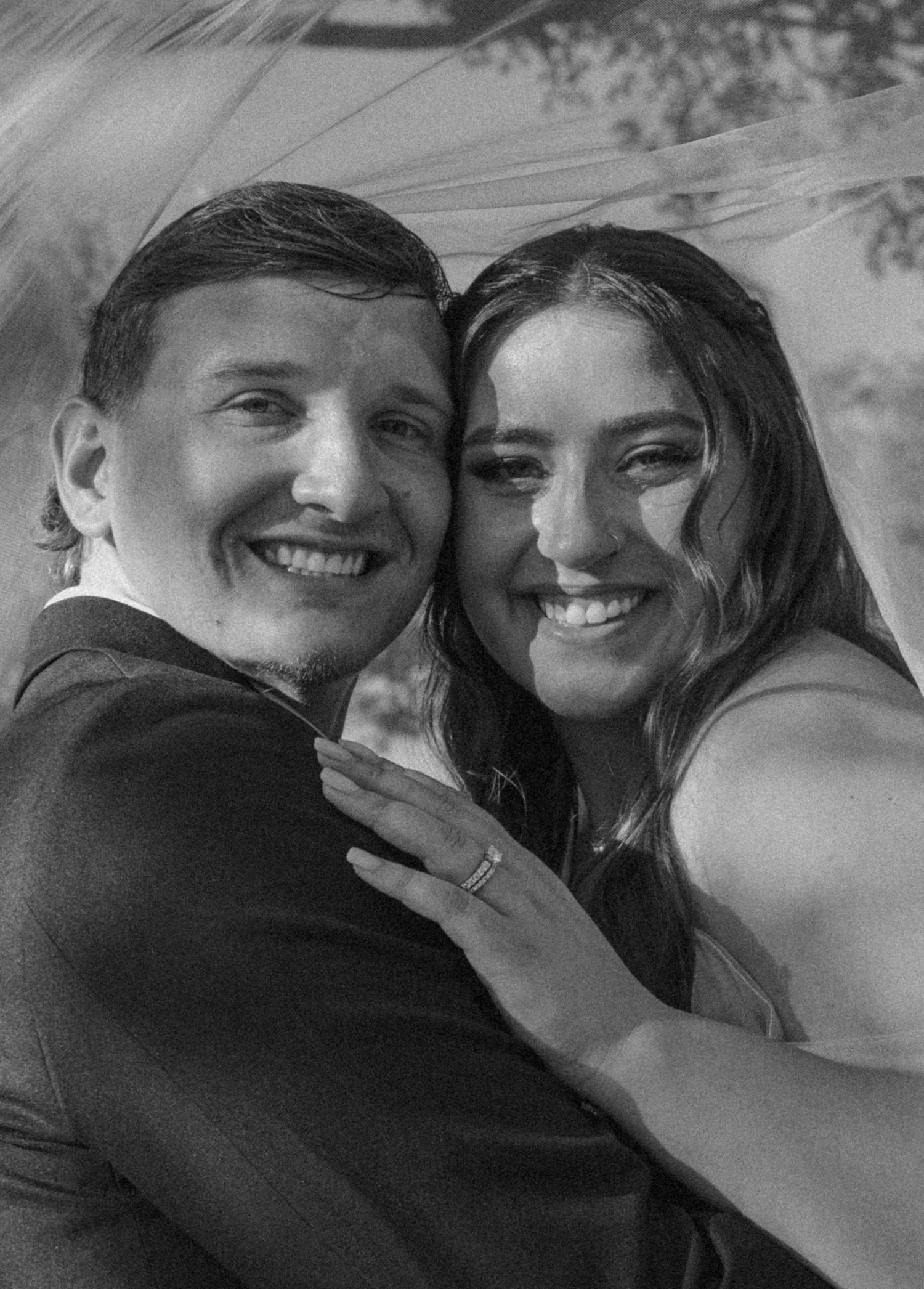 Black and white photo of a smiling couple embracing, woman wearing a ring, likely at a wedding celebration.