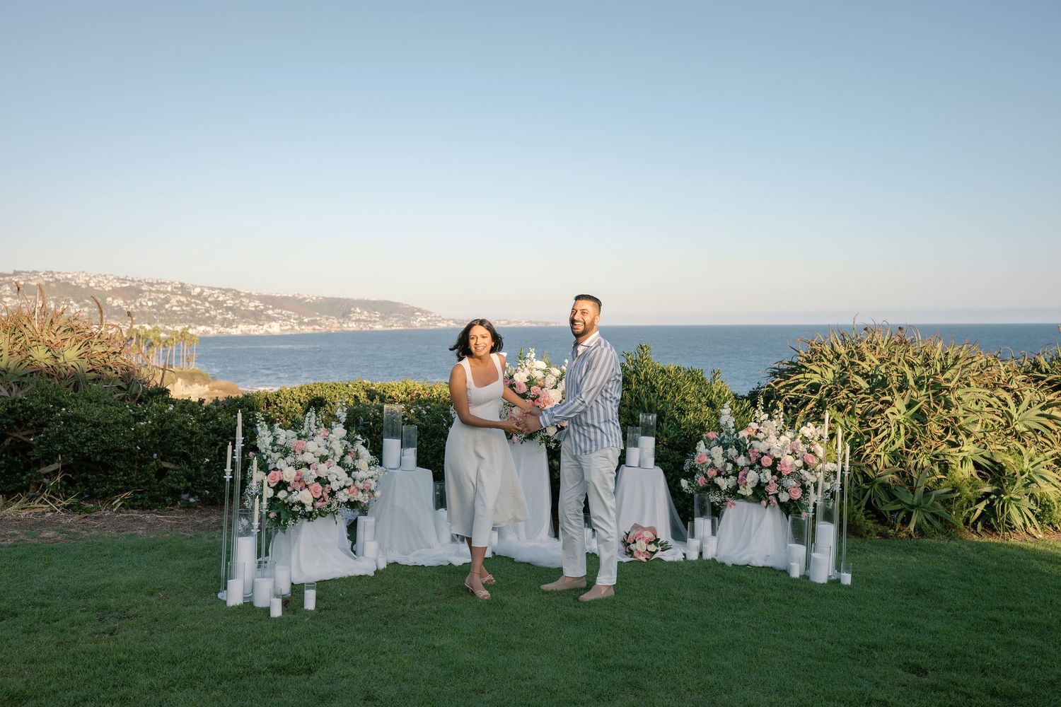 Couple holds bouquet at oceanview wedding ceremony with floral arrangements and candles on green lawn at sunset.