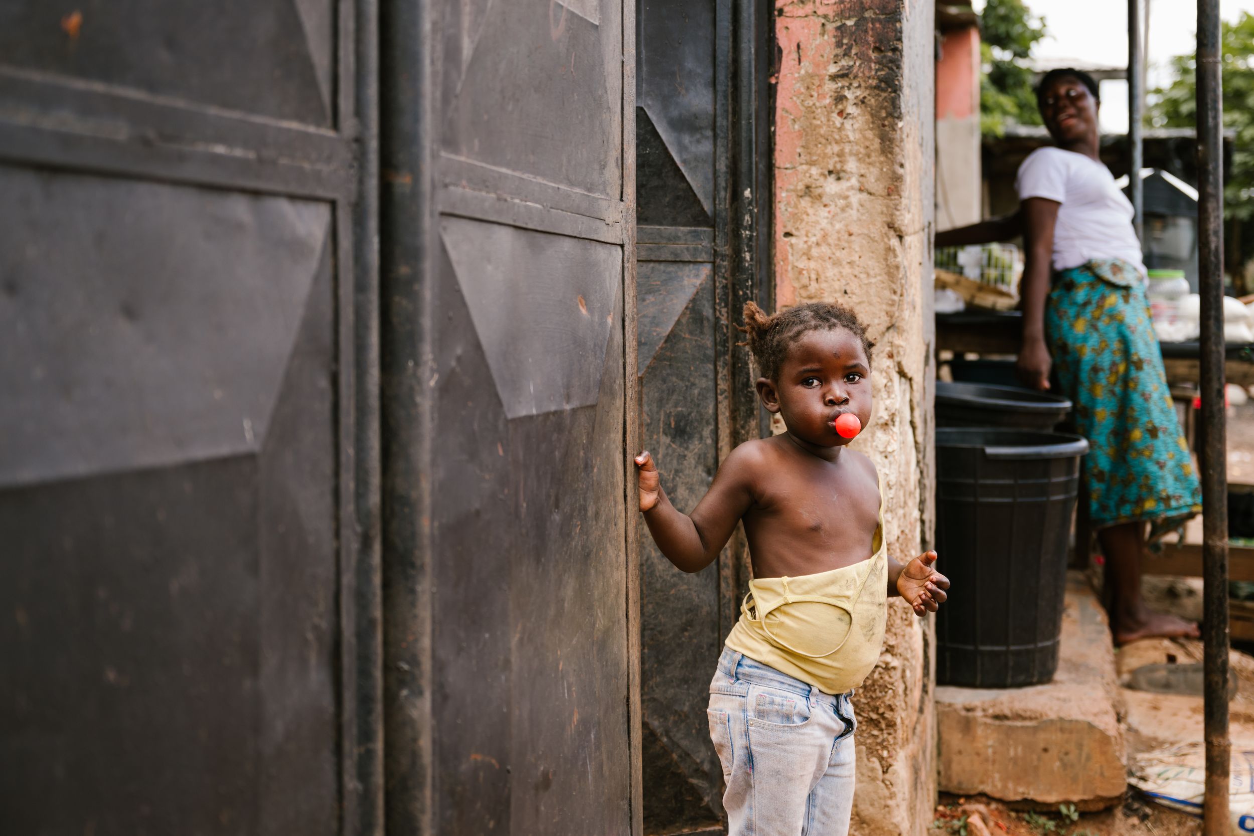 Young child standing in doorway of building in West Africa, with woman in colorful dress in background.