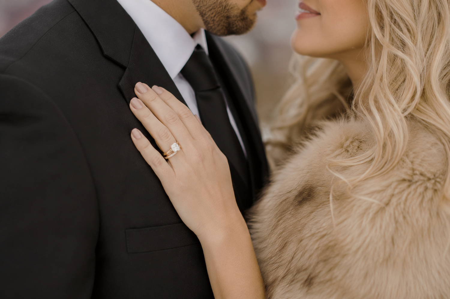 Couple in winter wedding attire, woman in fur stole with diamond ring, embracing against snowy backdrop.