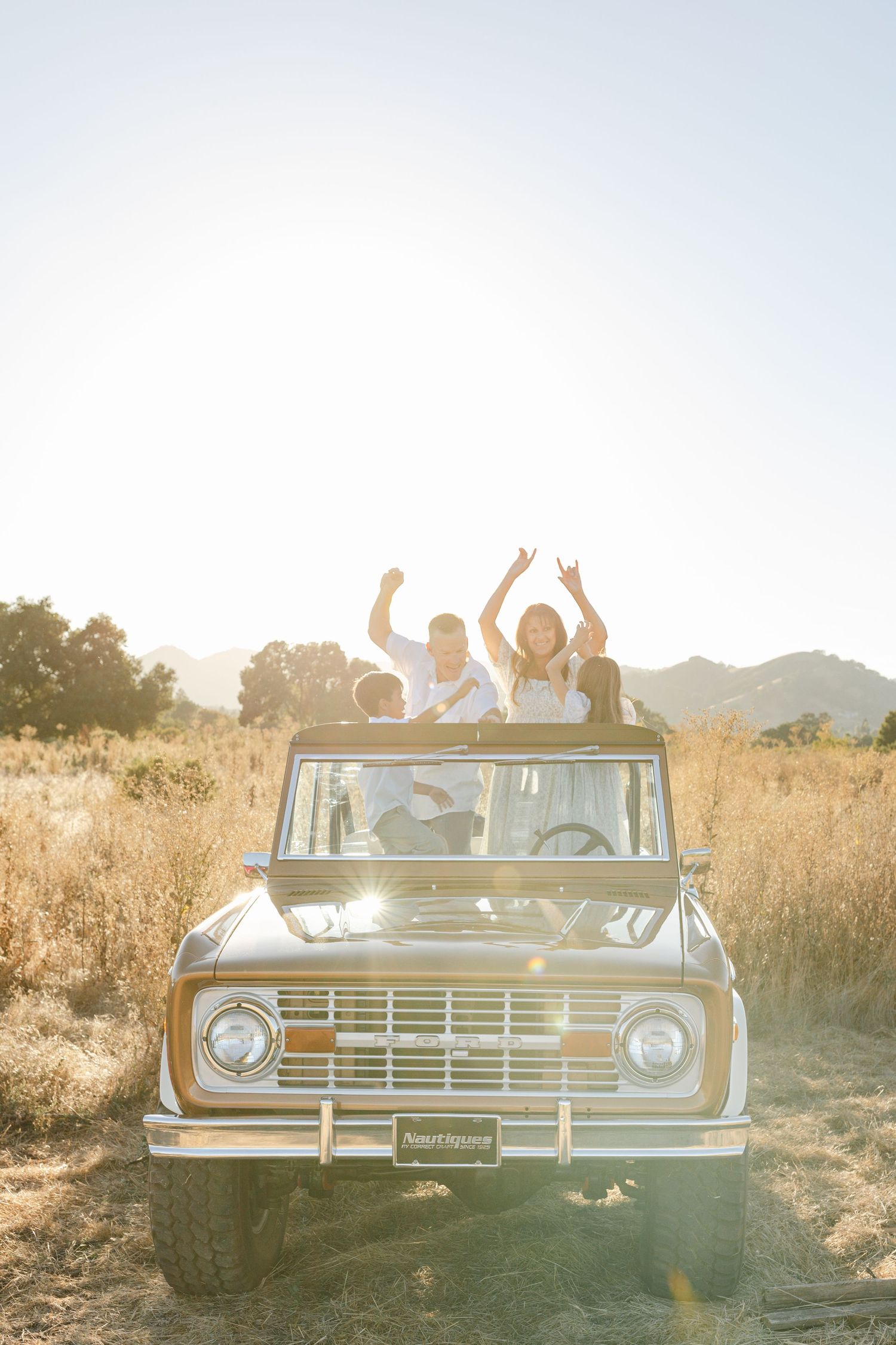 Group of friends joyfully riding in a vintage Ford Bronco through golden fields with mountains in the background.