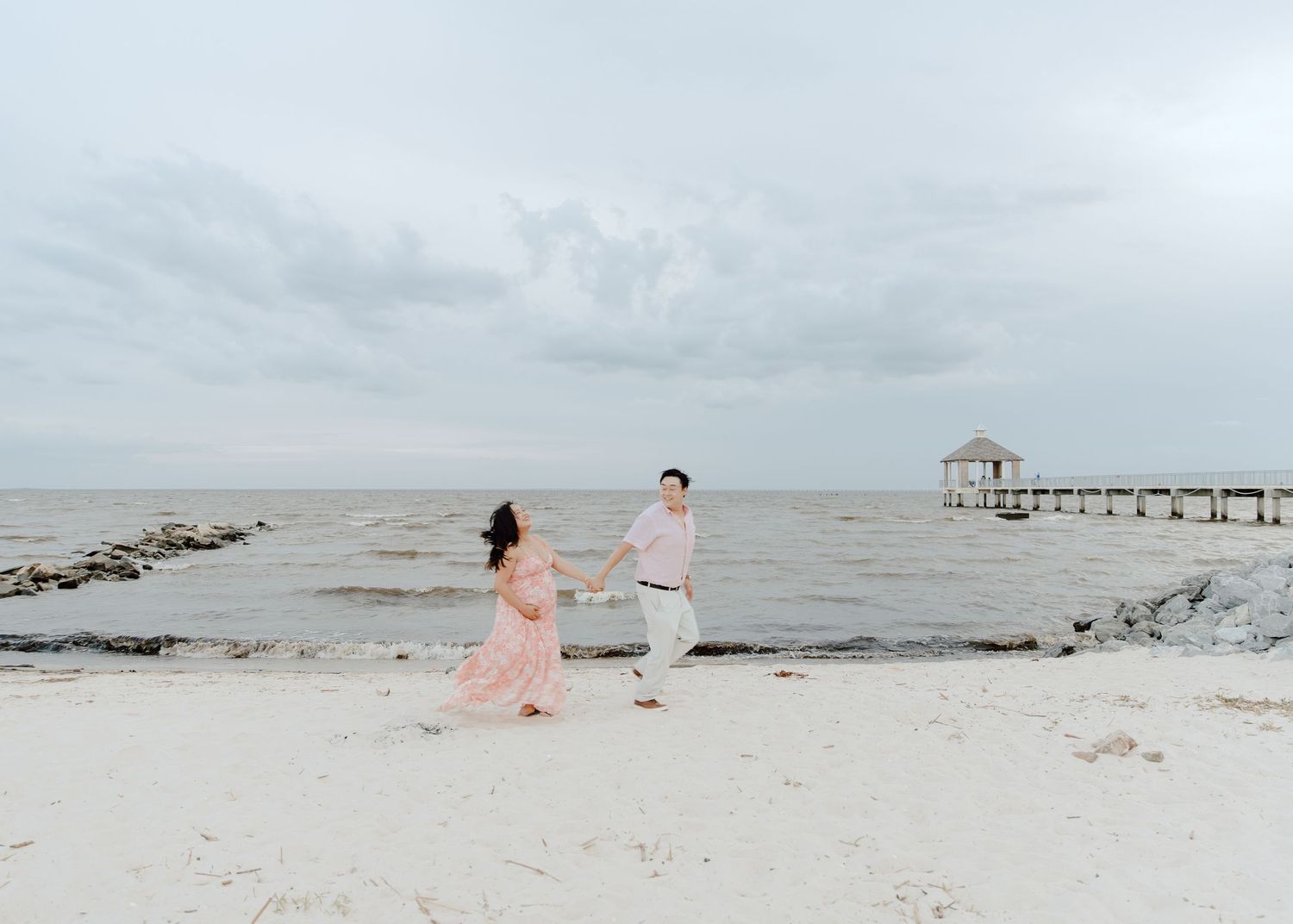 Couple holding hands walking on a sandy beach with a pier and calm ocean in the background under cloudy skies.
