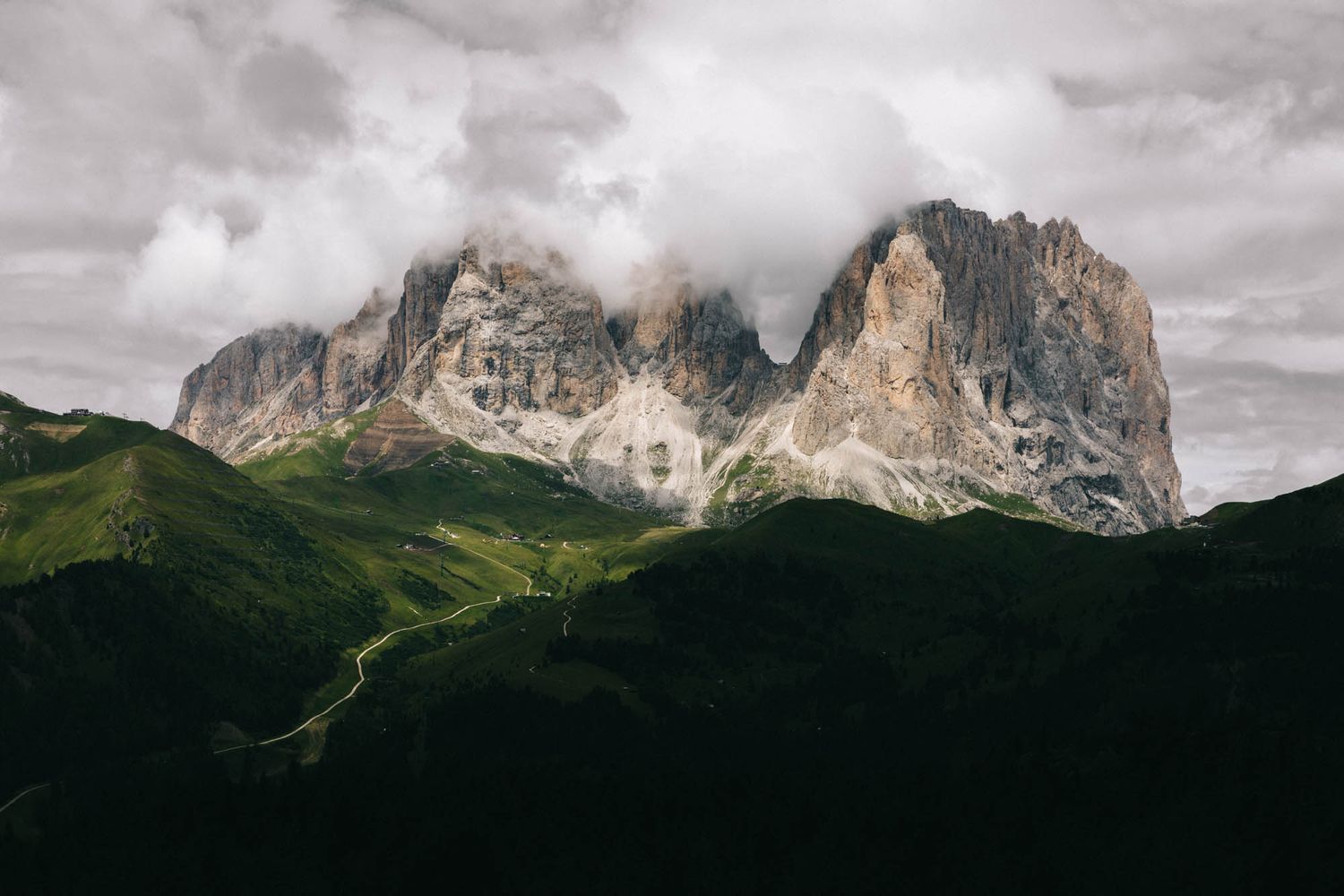 Dramatic alpine landscape featuring rocky Dolomite peaks rising above green rolling hills under stormy cloudy skies.