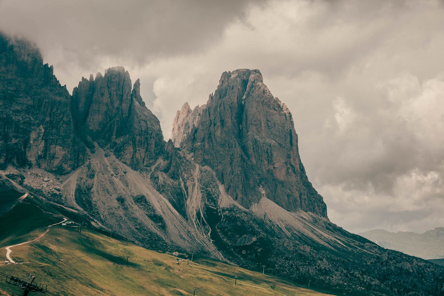 Dramatic rocky mountain peaks rise under stormy clouds in the Dolomites, with winding roads and green slopes below.