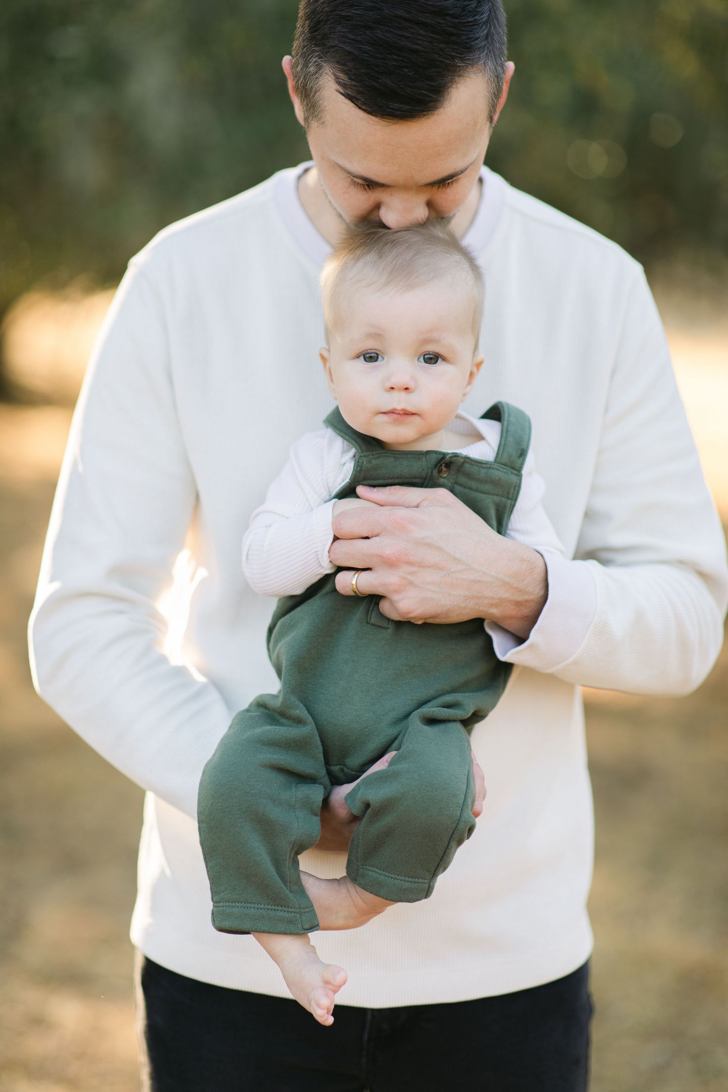 Father in white sweatshirt tenderly holding baby in green romper outdoors in natural setting.