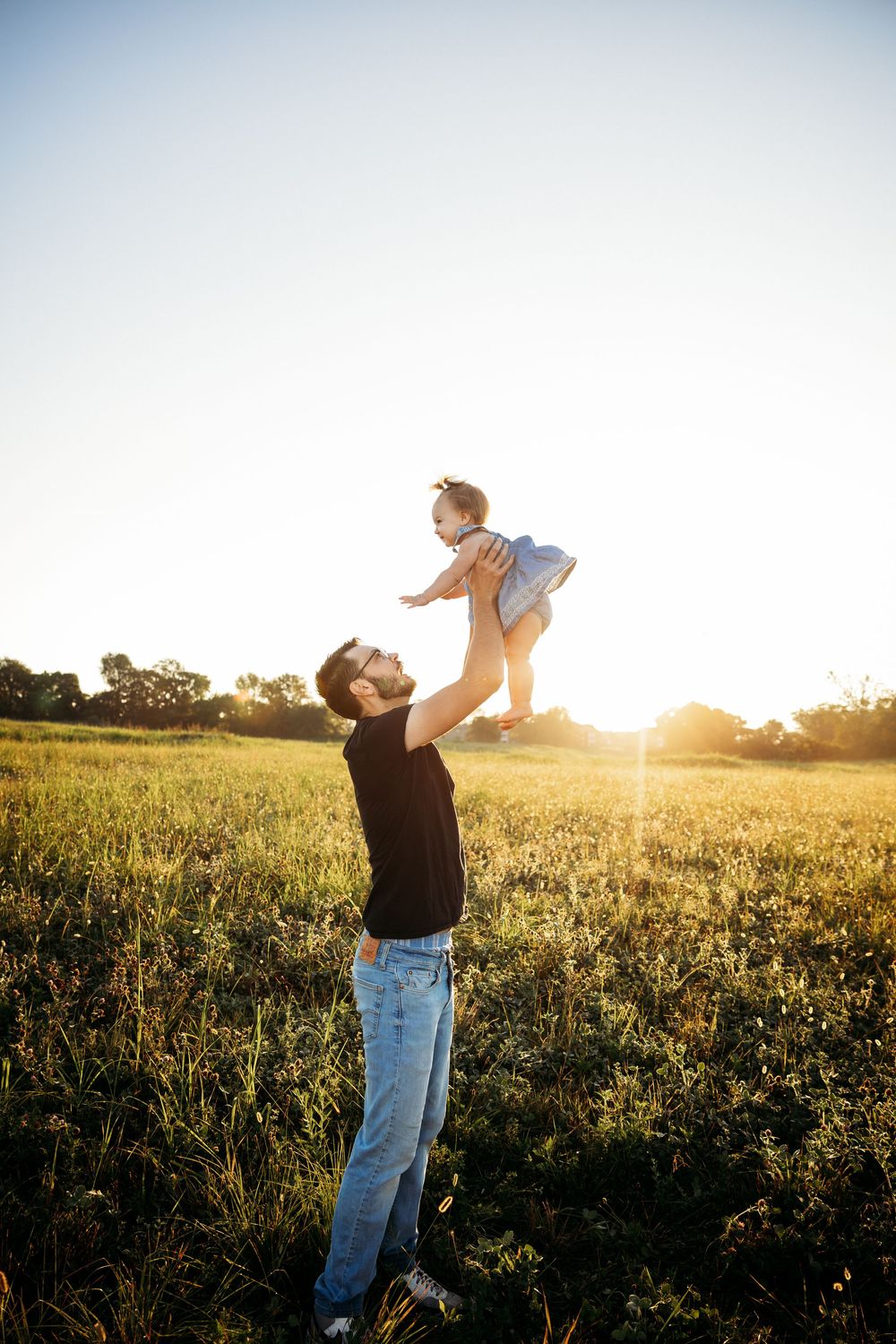 Father joyfully lifting baby daughter high in sunny green field at golden hour sunset.