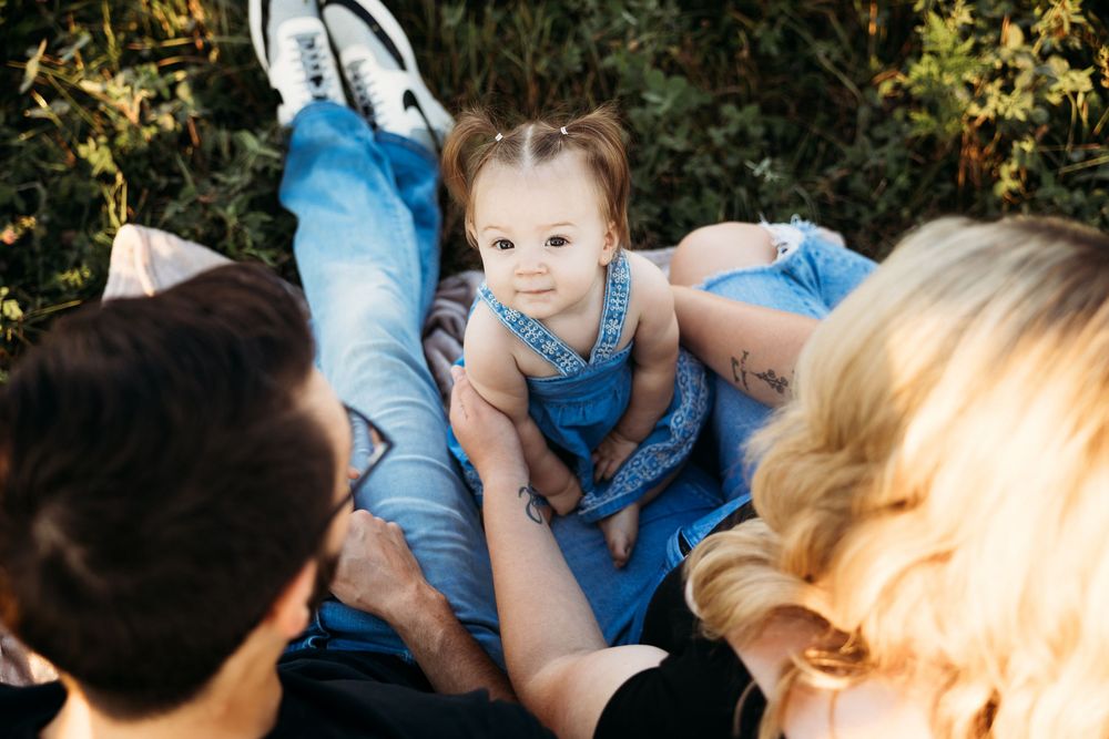 A smiling baby in blue overalls sits between two parents outdoors, surrounded by lush green foliage.